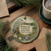 A decorative ornament featuring a scenic landscape of William B. Umstead State Park in North Carolina, with pine trees and a stream, is displayed on a wooden surface alongside a cup of coffee and some papers.