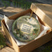A decorative ornament in a box, featuring a scenic landscape with a river, trees, and wildlife.