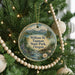 A round ornament hanging on a Christmas tree, featuring a scenic landscape with mountains, a river, and a bridge.
