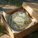 A circular ornament in a brown box, featuring a scenic landscape with a river, mountains, and a bridge.