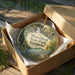 A decorative ornament in a wooden box, featuring a scenic image of Woodford State Park, Vermont.