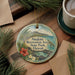 A round ornament with a tropical beach scene and text, hanging on a wooden surface next to a cup of coffee and some pine branches.