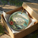 A round ornament in a box, featuring a tropical landscape with a mountain, palm trees, and a flower, along with the text "Auhukini Recreation Area State Park, Hawaii".