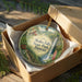 A circular ornament with a scenic image of Akaka Falls State Park in Hawaii, displayed in a wooden box with a gold string.