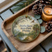 A wooden tray with a decorative ornament featuring a scenic view of Alafia River State Park, Florida, along with pine cones and a lit candle.