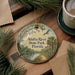 A round ornament with a painting of a river and wildlife, including a bird, hangs on a wooden surface.