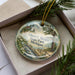 A decorative ornament in a box, featuring a scenic landscape with a lighthouse and a river.