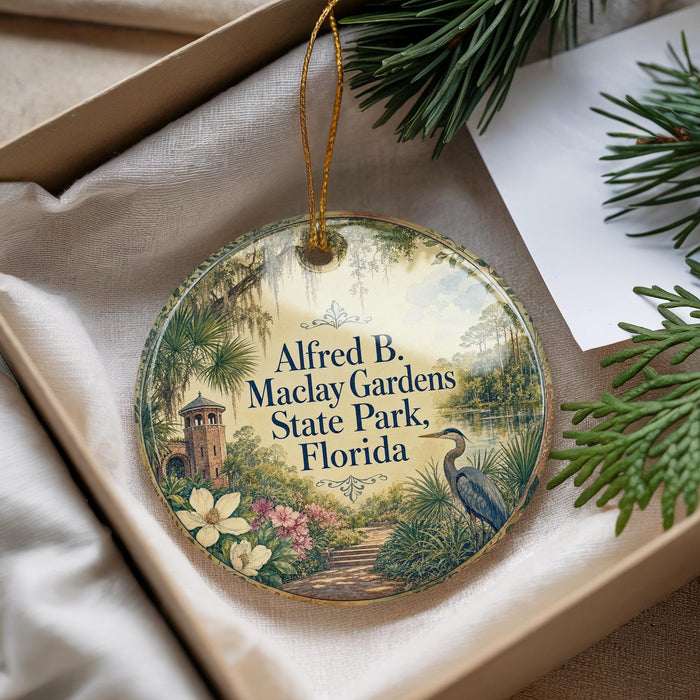 A round ornament with a tropical garden scene, featuring a bird, flowers, and a path, is displayed in a box with pine branches.
