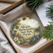 A round ornament with a tropical garden scene, featuring a bird, flowers, and a path, is displayed in a box with pine branches.