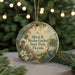 A round ornament with a floral design and text, hanging on a string, with a blurred background of greenery and lights.