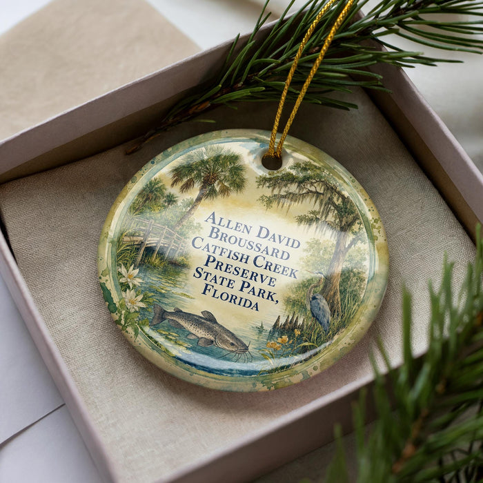 A decorative ornament with a painting of a fish and palm trees, placed in a box with a pine branch.