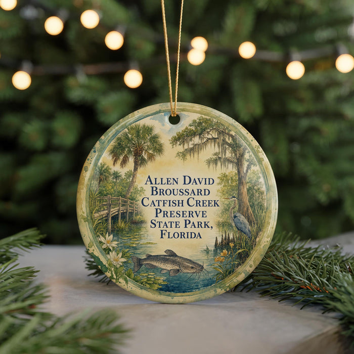 A round ornament with a painting of a pond and trees, featuring text that reads "Allen David Broussard, Catfish Creek, Preserve, State Park, Florida".