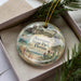 A decorative ornament with a beach scene and a bird is displayed in a box, surrounded by pine branches.