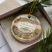A decorative ornament in a box, featuring a desert landscape with mountains, cacti, and a bighorn sheep.