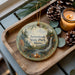 A decorative ornament with a scenic landscape of a waterfall and pine trees, featuring the text "Aroostook State Park, Maine". The ornament is placed on a wooden tray, with pine cones and a lit candle nearby.