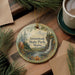 A decorative ornament with a scenic landscape of a waterfall and pine trees, featuring the text "Aroostook State Park, Maine". The ornament is placed on a wooden surface, surrounded by pine branches and a cup of coffee.
