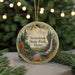 A circular ornament with a scenic landscape of a waterfall, pine trees, and autumn foliage, hanging in front of a blurred background with Christmas lights.