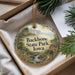 A decorative ornament with a scenic landscape of Backbone State Park, Iowa, featuring a waterfall and a house, is displayed in a box with pine branches.