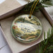 A circular ornament with a landscape scene and text, placed in a box with pine branches.