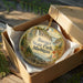 An oval ornament in a brown box, featuring a scenic landscape with a waterfall and trees, with the text "Baraga State Park, Michigan" printed on it.