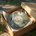 A round ornament in a box, featuring a scenic image of a waterfall surrounded by trees and greenery, with the text "Bash Bish Falls State Park Massachusetts" printed on it.
