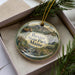 A decorative ornament in a box, featuring a scenic landscape with a moose, mountains, and a waterfall.