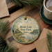 A round ornament with a scenic lake and park scene, featuring a bird and flowers, is displayed on a wooden surface alongside a cup of coffee and some pine branches.
