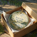 A round ornament in a box, featuring a scenic landscape with a lake, trees, and birds, with the text "Beets Lake State Park, Iowa" printed on it.