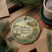 A round ornament with a painting of a house and trees, surrounded by greenery, is displayed on a wooden surface alongside a cup of coffee and some papers.