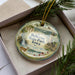 A decorative ornament in a box, featuring a scenic landscape with a waterfall, a castle, and a deer.