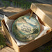 A round ornament in a box, featuring a scenic landscape with a lake and mountains, is displayed on a wooden surface.
