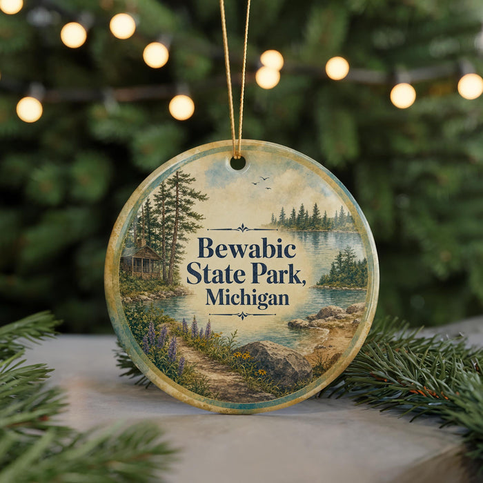 A round ornament with a painting of a scenic landscape, featuring a lake, pine trees, and a cabin, hanging from a string.