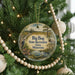 A round ornament hanging on a Christmas tree, featuring a scenic landscape with a moose, pine trees, and a path leading to a lake.