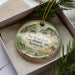 A decorative ornament in a box, featuring a scenic landscape with a barn, trees, and a river.