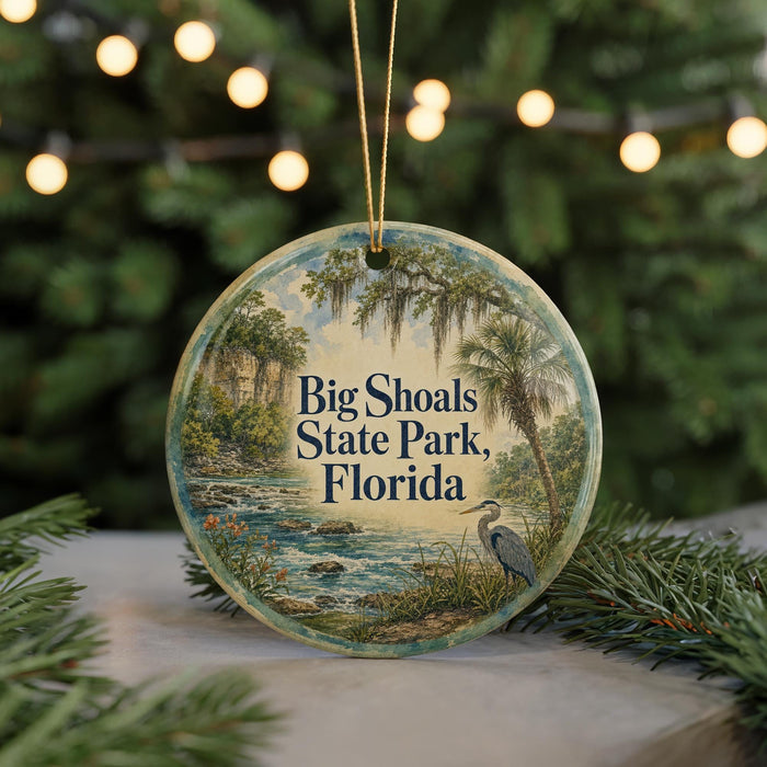 A round ornament with a painting of Big Shoals State Park, Florida, featuring a bird and a river, is displayed on a table with a blurred background of a Christmas tree.