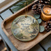A wooden tray with a decorative ornament featuring a scenic image of Big Talbot Island, Florida, surrounded by pine cones and a lit candle.