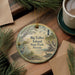 A decorative ornament featuring a scenic landscape with a lighthouse, birds, and palm trees, placed on a wooden surface alongside a cup of coffee and some pine branches.