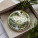 A decorative ornament in a box, featuring a scenic landscape with a waterfall and forest.