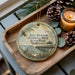 A round ornament with a scenic image of the Bill Burton Fishing Pier in Maryland, featuring a pier, boats, and a bird, is displayed on a wooden tray alongside pine cones and a lit candle.