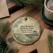 A round ornament with a painting of a pier and a bird, surrounded by pine branches and a cup of coffee on a wooden surface.