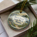 A decorative ornament in a box, featuring a scenic landscape with a body of water, trees, and a path.