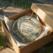 A decorative ornament in a box, featuring a scenic landscape with mountains, waterfalls, and greenery.