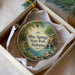 A decorative ornament in a white box, featuring a scenic landscape with a river, a bridge, and a bird perched on a rock.