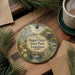 A round ornament with a painting of a scenic landscape hangs on a wooden surface, surrounded by pine branches and a cup of coffee.