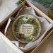 A decorative ornament in a box, featuring a scenic landscape with a river, trees, and a building.
