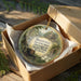 A circular ornament with a scenic image of a historic site, displayed in a cardboard box with tissue paper.