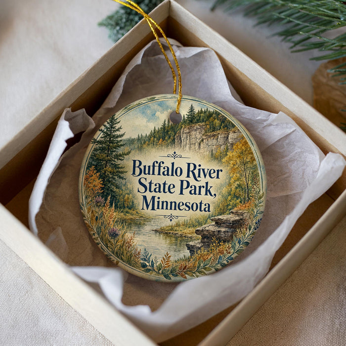 A decorative ornament in a box, featuring a scenic landscape with a river, trees, and mountains.