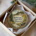 A decorative ornament in a box, featuring a scenic landscape with a river, trees, and mountains.