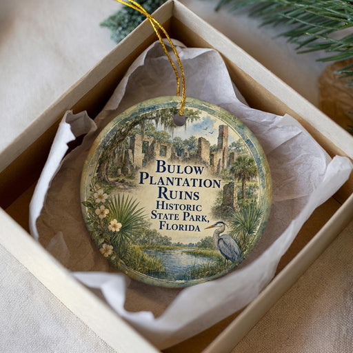 A circular ornament with a printed image of a tropical landscape, including palm trees, a bird, and a body of water, is displayed in a white box.