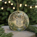 A circular ornament with a picture of a bird and palm trees, surrounded by greenery, hangs in front of a Christmas tree with lights.